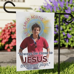 May include: A white garden flag with a photo of a young man with the text "ST. CARLO ACUTIS" above his head and the text "Always be close to JESUS, that's my plan." below his photo.