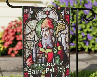 Puede incluir: Una bandera de jardín colorida con un diseño de vitral que representa a San Patricio sosteniendo un bastón. La bandera tiene el texto "Happy feast of Saint Patrick" en la parte inferior.