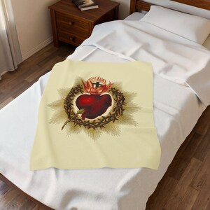 May include: A cream-colored throw blanket featuring a central graphic of a dark red Sacred Heart encircled by a brown crown of thorns, topped with a small black cross and surrounded by radiating golden light rays.