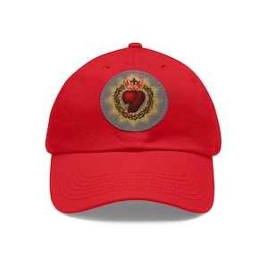 Peut inclure: Casquette de baseball rouge avec un patch circulaire représentant une image stylisée d'un cœur avec une couronne d'épines et une croix.