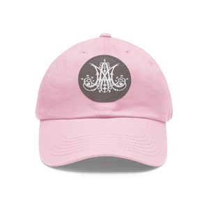 Peut inclure: Une casquette de baseball rose avec un patch en cuir marron orné d'un motif stylisé blanc. Le motif est un motif circulaire avec un motif central de trois lettres entrelacées.