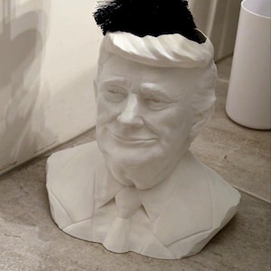 Op de afbeelding: Witte keramische toiletborstelhouder in de vorm van een buste van een persoon met een zwarte toiletborstel erin.