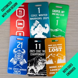 May include: Six different metal disc golf bag tags with various designs and text. The tags are all rectangular and have a hole at the top for attaching to a bag. The designs include a gorilla, a viking, a buffalo, a mountain range, and a set of icons for eating and sleeping. The text on the tags includes "CDGC Winter Warriors", "Casper, WY Disc Golf Club", "Not All Who Wander Are Lost", "2023 CDGC Tag Championship", and "Some Are Looking For It".