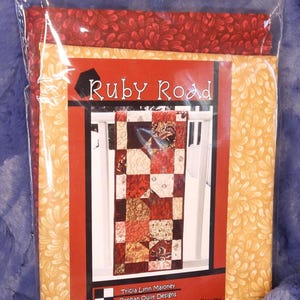 May include: A quilting pattern for a table runner called "Ruby Road" by Tricia Lynn Maloney for Villa Rosa Designs. The pattern features a red, brown, and cream patchwork design.