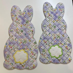 May include: Two quilted bunny-shaped placemats. The placemats are lavender with a floral pattern and a diamond stitch. Each bunny has a white cloud-shaped applique with a yellow or green border.