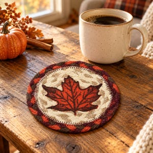 May include: A round, braided fabric coaster featuring a central cream-colored woven background with a prominent dark orange and brown maple leaf design, finished with a thick, multi-colored braided border in shades of red, orange, and brown.