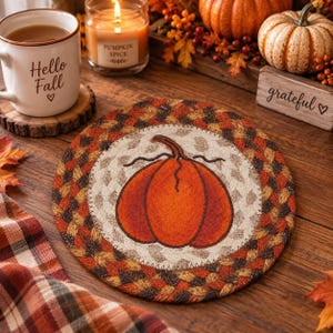 May include: A round, braided fabric coaster featuring a central cream-colored circle with a large, orange pumpkin illustration. The border consists of interwoven strips in shades of brown, orange, and tan, creating a textured, autumnal design.