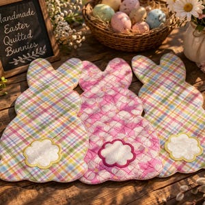 May include: Three handmade quilted bunny decorations in pastel colours. Two are plaid, one is pink floral. Each has a white cloud-shaped tail with a yellow border. A chalkboard sign reads "Handmade Easter Quilted Bunnies."