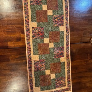 Peut inclure: Chemin de table matelass&eacute; rectangulaire avec un motif patchwork. Le chemin de table pr&eacute;sente des carr&eacute;s de tissu marron, vert et &agrave; motifs floraux, encadr&eacute;s par une bordure beige. La palette de couleurs comprend des nuances de marron, de vert et de beige.