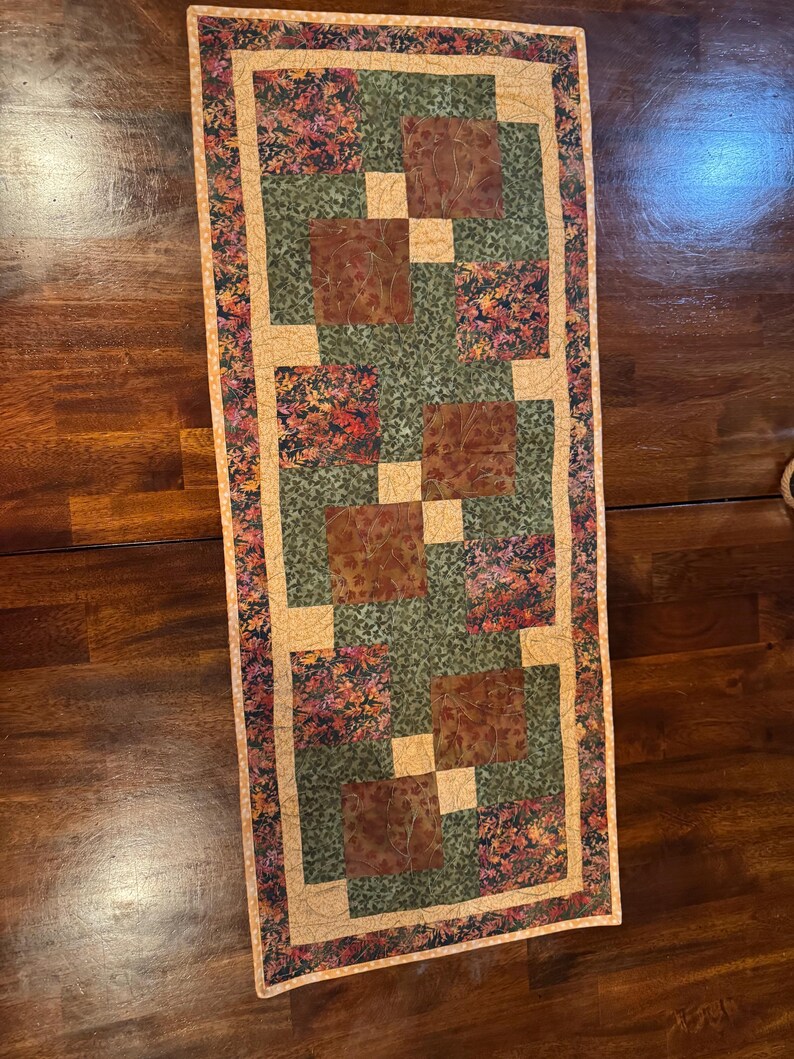 Peut inclure: Chemin de table matelass&eacute; rectangulaire avec un motif patchwork. Le chemin de table pr&eacute;sente des carr&eacute;s de tissu marron, vert et &agrave; motifs floraux, encadr&eacute;s par une bordure beige. La palette de couleurs comprend des nuances de marron, de vert et de beige.