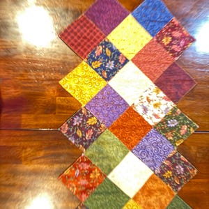 May include: A colorful, quilted table runner with a diamond pattern. The runner features squares of various fabrics in shades of red, yellow, blue, green, and purple. Some squares have floral or swirl patterns.