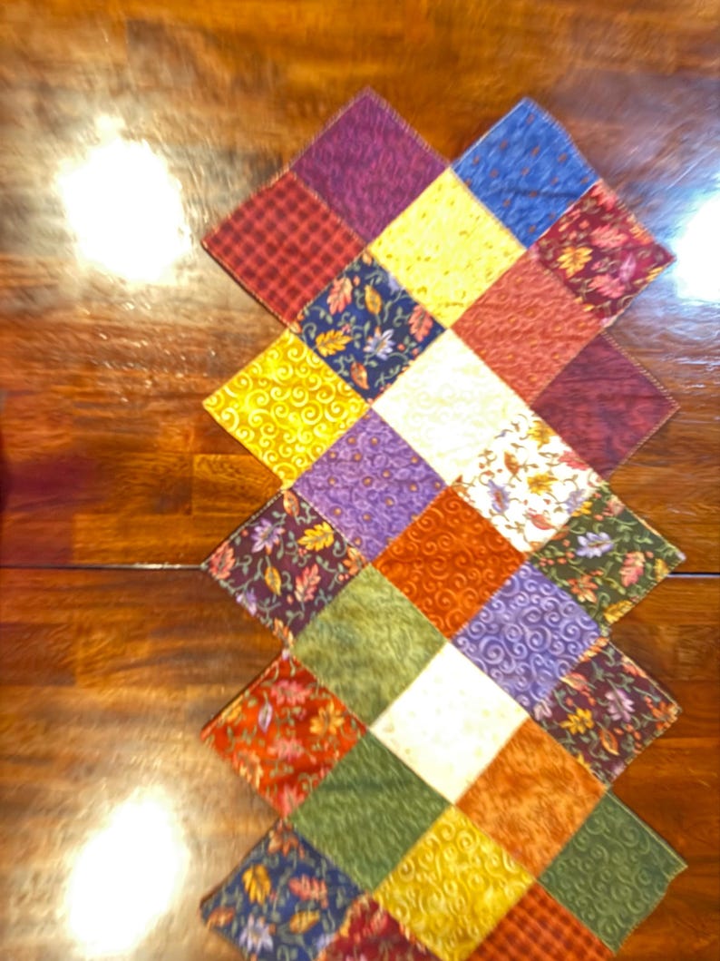 May include: A colorful, quilted table runner with a diamond pattern. The runner features squares of various fabrics in shades of red, yellow, blue, green, and purple. Some squares have floral or swirl patterns.