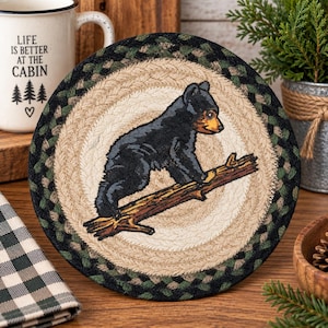 May include: A round braided coaster featuring a black bear cub climbing on a brown tree branch. The coaster has a central cream-colored circular area surrounded by a thick border of braided fabric in shades of dark green, black, and tan.