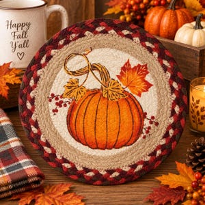May include: A round braided fabric mat featuring a central illustration of an orange pumpkin with a green stem, yellow leaves, and red berries. The mat has a cream-colored center surrounded by a thick border of braided red, brown, and tan fabric.