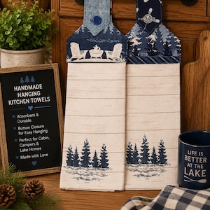 May include: Two hanging kitchen towels with button closures, featuring a light wood grain pattern and dark blue pine tree graphics. The top sections display campfire and paddle motifs in blue tones. A sign reads HANDMADE HANGING KITCHEN TOWELS.