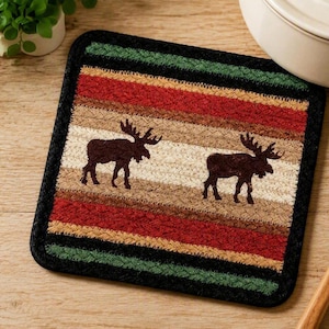 May include: A square braided fabric trivet featuring a horizontal striped pattern in black, green, red, tan, and brown. Two dark brown moose silhouettes are centered on the cream-colored middle stripe. The edges are finished with a thick black border.