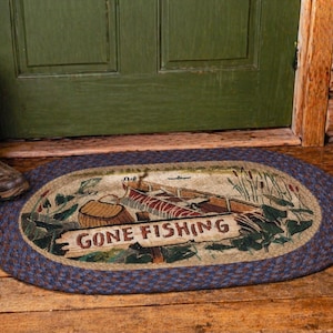 May include: Oval braided rug with a fishing theme. The rug features a boat, fishing gear, and the words "GONE FISHING". The border is a dark blue, and the rug is placed in front of a green door and brown boots.