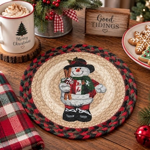 May include: A round braided jute placemat featuring a central illustration of a snowman wearing a black hat, a green and red scarf, and black boots. The placemat has a thick border of braided red, green, and black fibers surrounding the center.