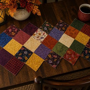 May include: A colorful patchwork table runner with a diamond pattern. The runner features squares of various fabrics in shades of blue, yellow, red, green, and purple. A red mug of coffee and a decorative lantern are also visible.