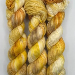 May include: Three skeins of yarn in shades of yellow, cream, and brown. The yarn is twisted into a skein shape, with the colors blending together. The yarn is likely for knitting or crocheting projects.