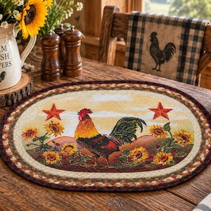 May include: An oval placemat featuring a central rooster standing among sunflowers and two red stars against a light background. The placemat has a dark, multi-coloured braided border. The design includes warm tones of yellow, orange, red, and brown.
