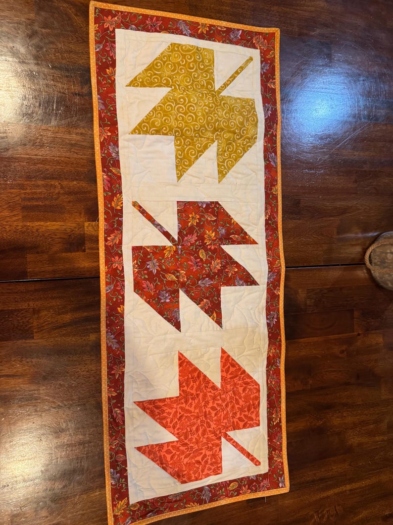 May include: A quilted table runner with three leaf designs in gold, red, and orange. The leaves are set against a cream background, with a floral border. The runner is approximately 30 cm wide and 91 cm long, suitable for a dining table.