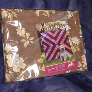 May include: A brown fabric with a floral pattern is packaged in a clear plastic bag. A quilting pattern for a quilt called "Birdie" is visible in the bag. The pattern is for a quilt with a purple and orange design. The pattern is from Running Doe Quilts for Villa Rose Designs.