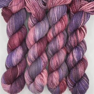 May include: Four skeins of hand-dyed yarn in shades of purple, pink, and burgundy. The yarn is twisted into a rope-like shape, showing color variations throughout. The skeins are arranged side by side, showcasing the rich, saturated colors.