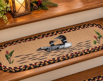 May include: An oval braided stair tread featuring a natural tan center with a black and red braided border. The center depicts two loons swimming on water with green and red floral accents on the left and right sides of the rug.