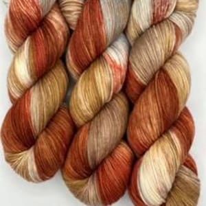 May include: Three skeins of hand-dyed yarn in a gradient of warm colors. The yarn transitions from a deep rust to shades of tan, cream, and grey. The skeins are tightly wound and displayed against a white background.