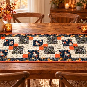 May include: A rectangular table runner featuring a patchwork design with alternating blocks of dark navy blue fabric with orange floral patterns and cream fabric with small floral prints, accented by solid orange squares and a thin orange border.