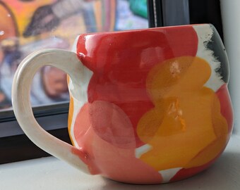 May include: A white ceramic mug with a red, orange, and pink abstract design. The mug has a rounded shape and a white handle.