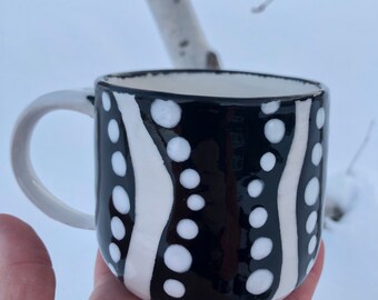 May include: A black and white ceramic mug with a wavy pattern of white stripes and white polka dots. The mug has a white handle and a white interior. The mug is held in a hand against a snowy background.