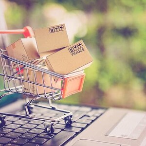Könnte beinhalten: Ein Miniatur-Einkaufswagen gefüllt mit Kartons, von denen einige mit "Fragile" beschriftet sind, steht auf einer Laptop-Tastatur. Die Kartons sind braun und haben schwarze Markierungen. Der Einkaufswagen ist silberfarben mit einem roten Griff.