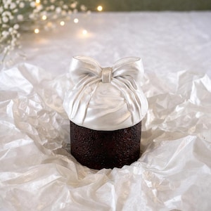 Peut inclure: Un gâteau cylindrique brun foncé est surmonté d'un nœud blanc satiné. Le gâteau repose sur du papier froissé blanc. L'arrière-plan est vert tendre avec des lumières floues et des fleurs blanches.