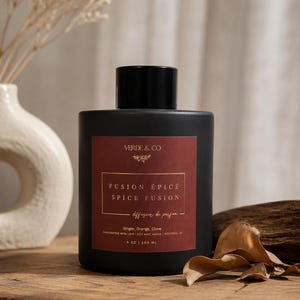 May include: A matte black cylindrical fragrance diffuser bottle with a dark red rectangular label. The label features the text VERDE & CO, FUSION ÉPICÉ SPICE FUSION, and lists Ginger, Orange, Clove, with a volume of 160 ml or 5 oz printed on it.