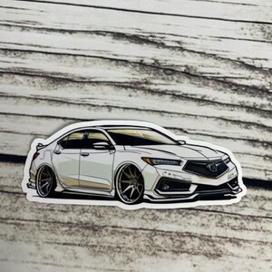 May include: A white car sticker featuring a detailed illustration of a white saloon car with black accents and gold wheels. The car has a black front grille with a silver emblem.
