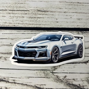 May include: A silver Chevrolet Camaro sports car sticker featuring a sleek aerodynamic design, a black front grille, sharp angular headlights, and dark multi-spoke wheels. The vehicle is depicted in a three-quarter view with a rear spoiler.