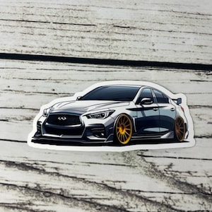 May include: A sticker featuring a white Infiniti sedan with a lowered stance, a front splitter, and bright gold multi-spoke wheels. The car is depicted in a stylized graphic illustration with dark grey shading and a small rear spoiler.