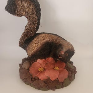 May include: A detailed figurine of a skunk with a curved tail, resting on a base with two red flowers and green leaves. The skunk is primarily brown and black, with a textured fur appearance. The base has a detailed, earthy design.