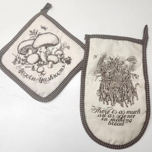 May include: A square pot holder and a rectangular oven mitt in off-white fabric with brown polka dot trim. The square piece features a black line drawing of mushrooms and the text Meadow Mushrooms. The mitt shows wheat stalks and the text There is as much art as science in making bread.