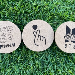 May include: Three round cork coasters with black printed designs. The first coaster has a heart and the text "BORAHAE II". The second coaster has a hand making the Korean heart sign. The third coaster has the text "BTS" and a group of cartoon characters.