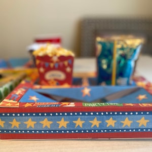 May include: A red and blue cardboard box with a window, decorated with stars and the text "Come One Come All" and "Party Time". The box is designed to hold snacks and drinks for a party.
