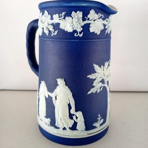 Puede incluir: Una jarra de cerámica azul y blanca con un diseño de vid blanca alrededor de la parte superior y una escena blanca de tres figuras y un árbol en la parte delantera.