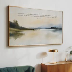 May include: A framed landscape art print depicting a tranquil lake scene in muted tones of blue, green, and beige. The artwork includes text at the top: "WHOEVER DRINKS THE WATER..." The print is presented in a light wood frame.