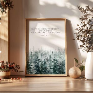 May include: A framed print with a watercolour forest scene in greens and blues. The print displays the text "GLORY TO GOD IN THE HIGHEST, AND ON EARTH, PEACE GOOD WILL TOWARD MEN!" from Luke 2:14. The frame is made of light-coloured wood.