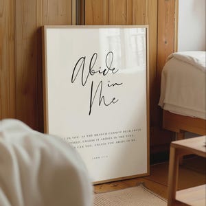 Könnte beinhalten: Ein rechteckiger Wandkunstdruck in einem hellen Holzrahmen mit dem schwarzen Text Abide in Me in einer großen Schreibschrift, gefolgt von kleinerem Serifentext, der Johannes 15,4 zitiert, auf einem schlichten, cremeweißen Hintergrund.