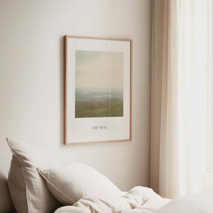 May include: A framed landscape art print with a light wood frame hangs on a white wall. The print features a muted color palette of green, beige, and blue, with the text "THE WAY" at the bottom. The artwork is displayed above a bed with white bedding.