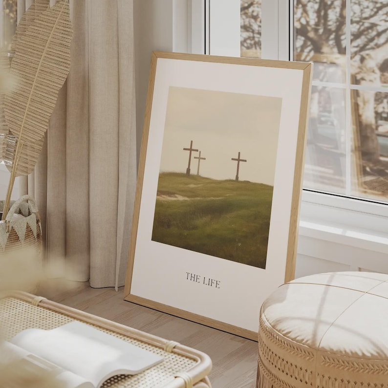 May include: Framed art print featuring three crosses on a grassy hill under a muted sky. The print is in a light wood frame and has the words "THE LIFE" at the bottom. The artwork is leaning against a wall near a window.