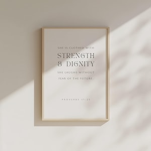 May include: A framed print with the text "SHE IS CLOTHED WITH STRENGTH & DIGNITY SHE LAUGHS WITHOUT FEAR OF THE FUTURE. PROVERBS 31:25" in a minimalist style. The frame is a light beige color, and the background is white.